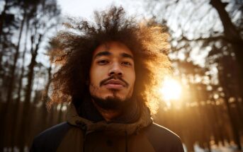 A man with curly hair and a beard stands outdoors in a winter forest at sunset, wearing a jacket. Sunlight shines through the trees behind him, creating a warm, glowing effect around his hair.