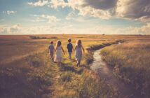 Four children walk along a grassy path beside a narrow stream in a sunlit field under a partly cloudy sky, creating a peaceful and nostalgic scene.