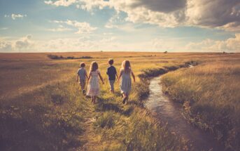 Four children walk along a grassy path beside a narrow stream in a sunlit field under a partly cloudy sky, creating a peaceful and nostalgic scene.