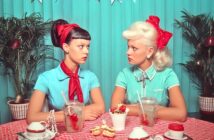 Two women with vintage hairstyles and red bows sit at a table with checkered tablecloth, drinks with cherries, and desserts, facing each other against a teal backdrop with plants and festive bunting.