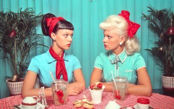 Two women with vintage hairstyles and red bows sit at a table with checkered tablecloth, drinks with cherries, and desserts, facing each other against a teal backdrop with plants and festive bunting.