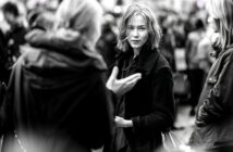 A young woman with short, messy hair stands in a crowded outdoor setting, looking directly at the camera while people around her talk and move. The image is in black and white.