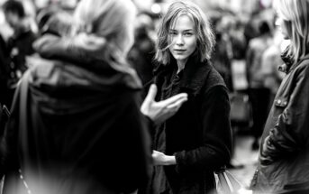 A young woman with short, messy hair stands in a crowded outdoor setting, looking directly at the camera while people around her talk and move. The image is in black and white.