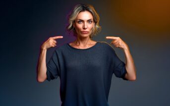 A woman with short blonde hair and a serious expression stands against a dark background, pointing at herself with both hands. She wears a loose dark gray top.