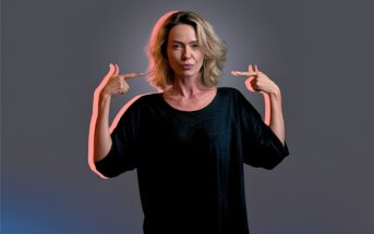 A woman with shoulder-length blonde hair in a black top stands against a gray background, confidently pointing both index fingers at herself.