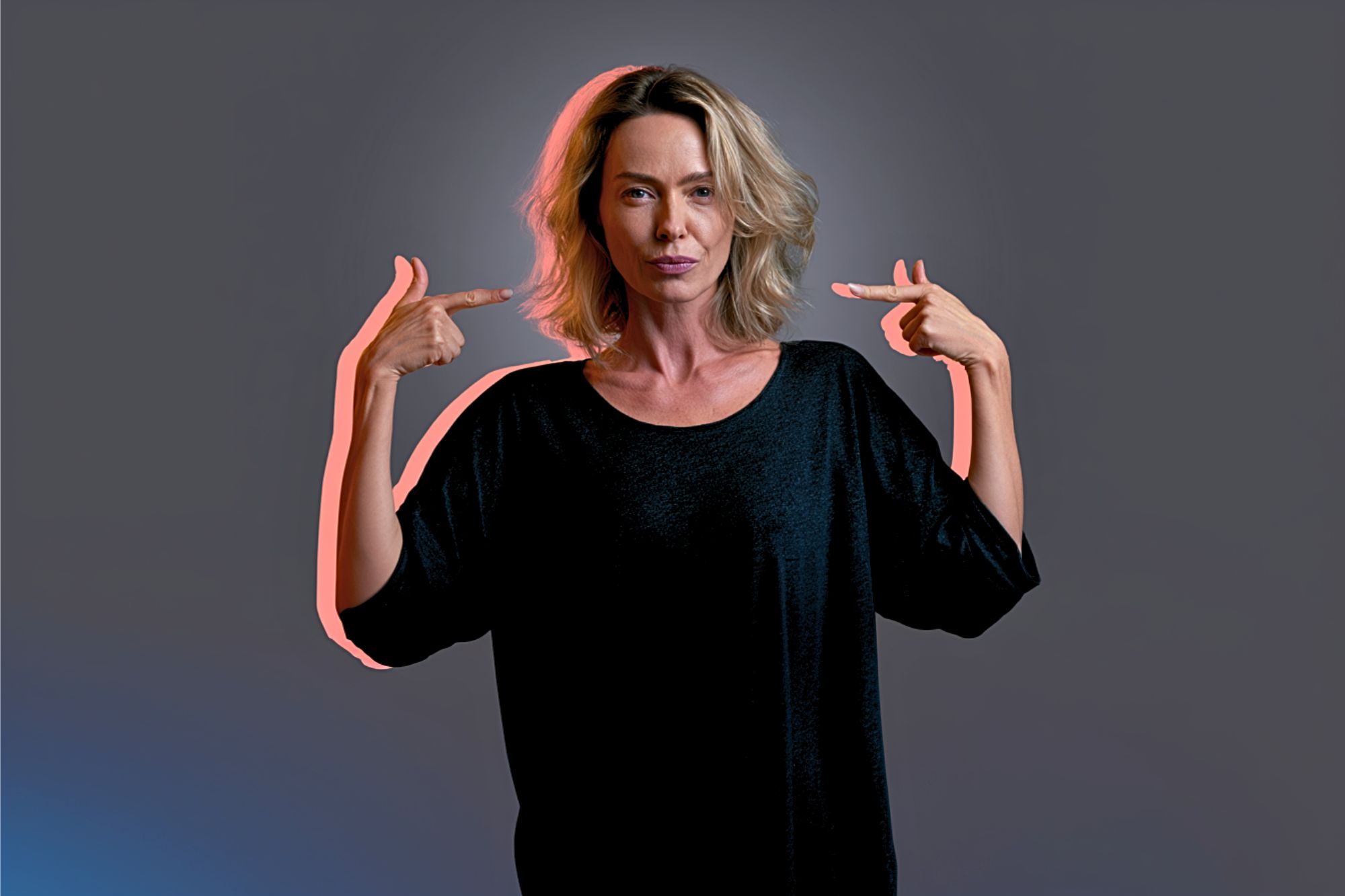 A woman with shoulder-length blonde hair in a black top stands against a gray background, confidently pointing both index fingers at herself.