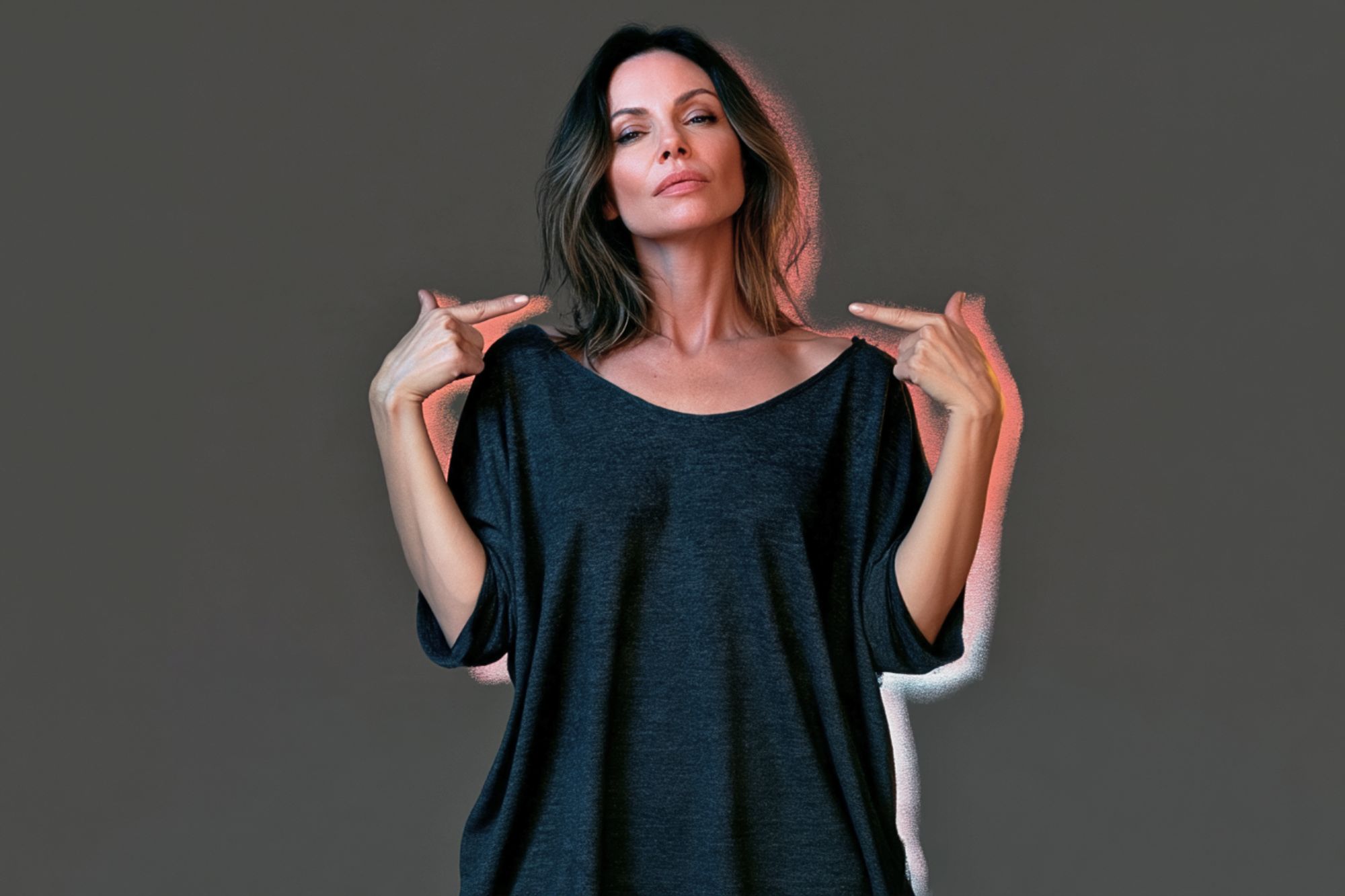 A woman in a dark grey oversized top points to her shoulders with both hands.