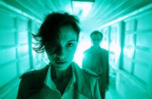 A woman with short dark hair stands in sharp focus under green lighting in a narrow hallway, looking directly at the camera; a blurred man stands behind her in the background.