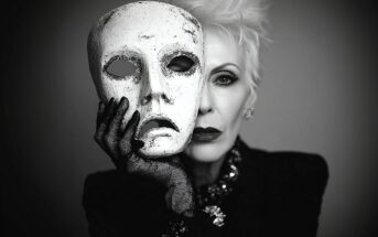 A woman with short, white hair and dramatic makeup holds a white, expressionless mask in front of her face, partially obscuring it. She wears dark clothing, lace gloves, and bold jewelry. The image is in black and white.