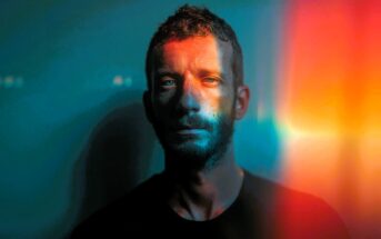 A man with short hair and a beard stands against a blue background, illuminated by colorful orange and yellow light streaks crossing his face, creating a dramatic, artistic effect.