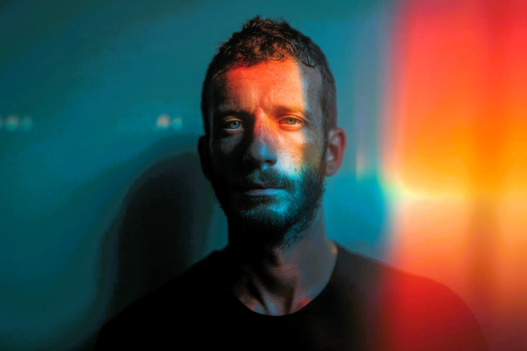 A man with short hair and a beard stands against a blue background, illuminated by colorful orange and yellow light streaks crossing his face, creating a dramatic, artistic effect.