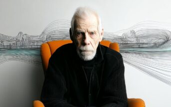An elderly man with white hair and a beard sits in an orange chair, wearing a black jacket. Behind him, abstract black lines and wavy patterns are drawn on a white wall.