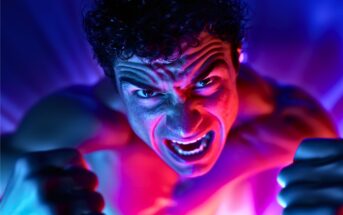 A man with curly hair screams intensely, clenching his fists. He is illuminated by dramatic blue and pink lighting, creating a sense of anger and energy. The background is blurred with vibrant, radiating colors.