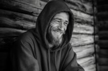 A bearded man in a hooded sweatshirt smiles warmly while leaning against a weathered wooden wall. The photo is in black and white, highlighting the texture of the wood and the man's joyful expression.