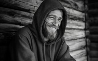 A bearded man in a hooded sweatshirt smiles warmly while leaning against a weathered wooden wall. The photo is in black and white, highlighting the texture of the wood and the man's joyful expression.