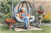 A woman with white hair in a blue dress sits relaxed on a cushioned hanging chair in a garden, surrounded by flowers, greenery, and a stone wall.