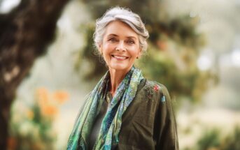 An older woman with gray hair smiles warmly, standing outdoors in front of blurred greenery. She wears a green top and a colorful scarf, radiating a peaceful and content demeanor.