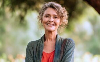 Smiling older woman with curly gray hair stands outdoors in a green, leafy setting, wearing a red blouse and a green shawl.
