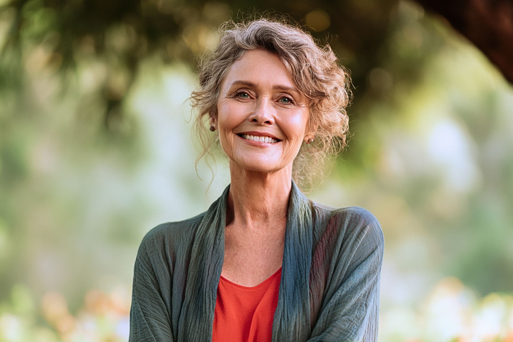 Smiling older woman with curly gray hair stands outdoors in a green, leafy setting, wearing a red blouse and a green shawl.