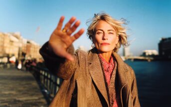 A woman with blonde hair and a brown coat stands on a riverside walkway, facing the camera with her hand outstretched, partially covering her face. The background shows water, buildings, and a clear blue sky.