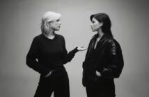 Two women stand facing each other against a plain background. One, with light hair, gestures with her hand while speaking; the other, with dark hair and a leather jacket, listens calmly with her hands in her pockets.