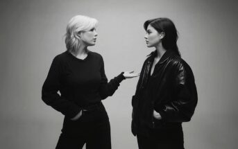 Two women stand facing each other against a plain background. One, with light hair, gestures with her hand while speaking; the other, with dark hair and a leather jacket, listens calmly with her hands in her pockets.