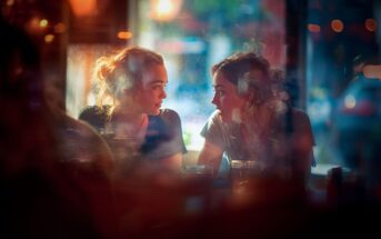 Two young women sit closely at a dimly lit café, engaged in an animated conversation. Warm light and reflections from the window create a soft, colorful atmosphere around them, emphasizing their connection.