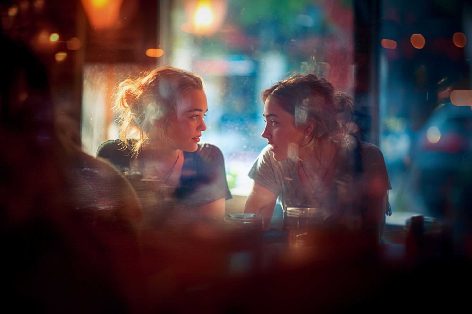 Two young women sit closely at a dimly lit café, engaged in an animated conversation. Warm light and reflections from the window create a soft, colorful atmosphere around them, emphasizing their connection.