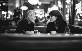 Two women sit across from each other at a café table, engaged in conversation. Both have cups in front of them. The background is warmly lit with blurred lights, creating a cozy and intimate atmosphere.