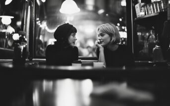 Two women sit across from each other at a café table at night, engaged in conversation. The scene is warmly lit with string lights and shelves in the background, creating a cozy, intimate atmosphere. The image is in black and white.