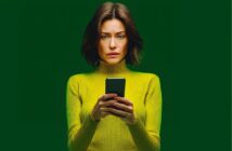 A woman with shoulder-length brown hair wearing a yellow-green turtleneck sweater holds a smartphone and looks at the screen with a concerned expression against a solid green background.