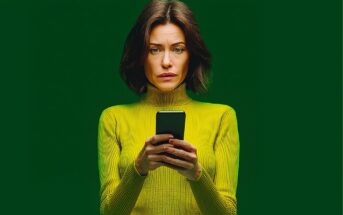 A woman with shoulder-length brown hair wearing a yellow-green turtleneck sweater holds a smartphone and looks at the screen with a concerned expression against a solid green background.