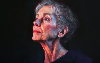 A mature woman with short gray hair gazes upward in soft, colorful lighting against a dark background, her expression thoughtful and serene. She wears a simple, dark top and small stud earrings.