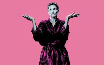 A woman in a robe stands against a pink background, raising both hands with a confused or uncertain expression.