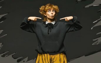 A woman with curly hair wearing a black sweater and yellow pants stands against a dark background, making a playful face and pointing at herself with both hands. The image has a cartoon-like effect.