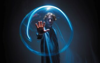 A person in a dark room extends their hand forward, surrounded by a glowing blue light ring, creating a mysterious and futuristic atmosphere.