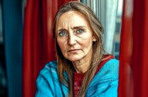 A woman with long brown hair and blue eyes, wearing a blue sweater and red top, sits indoors by a window with red curtains, looking directly at the camera with a serious expression.