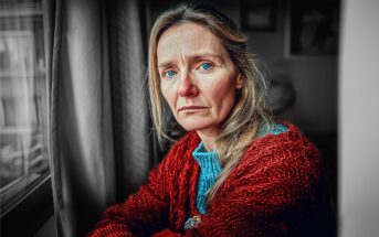 A woman with light hair and blue eyes, wearing a red sweater, sits by a window looking out with a serious, contemplative expression. The photo is mostly black and white except for her face and clothing, which are in color.