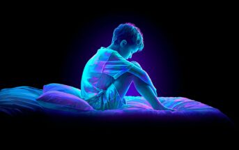 A young child sits alone on a bed in a dark room, illuminated by neon blue and purple light, with their knees drawn to their chest and head bowed, suggesting sadness or contemplation.
