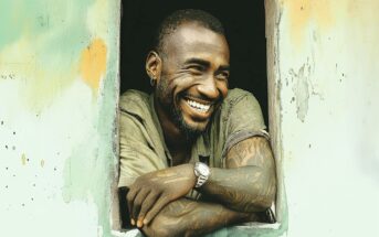 A smiling man with tattoos on his arms leans on a windowsill, wearing a watch and a short-sleeved shirt. The window frame is weathered and part of a wall with faded, peeling paint in green and orange tones.