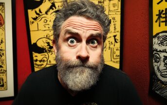A bearded man with wide eyes and raised eyebrows stares directly at the camera, standing in front of colorful cartoon-style artwork on a red wall.