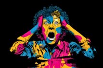 A person with curly hair holds their head and screams in shock. Their face and clothes are painted in bold, vibrant blue, yellow, and pink colors against a black background.