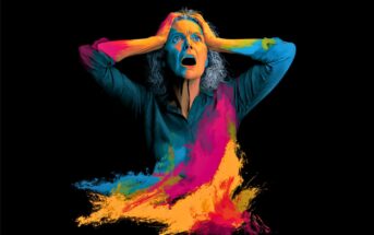 A woman with gray hair holds her head and appears distressed. Her face and body are dramatically painted with bright blue, yellow, pink, and orange colors on a black background.