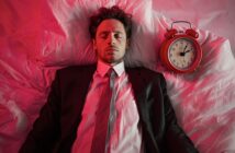 A man in a suit lies on a bed with his eyes closed, next to a red alarm clock. The scene is lit with pinkish-red light, creating a surreal atmosphere.