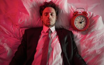 A man in a suit lies on a bed with his eyes closed, next to a red alarm clock. The scene is lit with pinkish-red light, creating a surreal atmosphere.