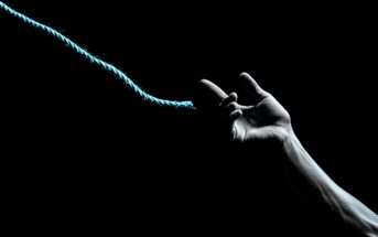 A dimly lit hand reaches out, fingers hooked around a blue rope against a black background, suggesting a moment of grasping or letting go.