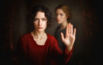 A woman with short dark hair and a serious expression stands in focus with her hand raised, palm facing forward. Another woman with lighter hair is blurred in the background. The scene is softly lit and dramatic.