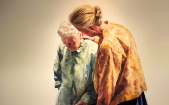 Two older women stand together; one in a green blouse looks down with a sad expression, while the other, in a yellow floral shirt, leans in to comfort her with a gentle hand on her shoulder against a plain background.