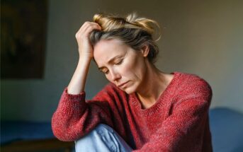 A woman wearing a red sweater sits with her hand on her head, eyes closed, appearing sad or stressed in a softly lit room.