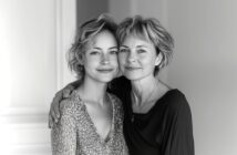 Two women, one younger and one older, stand close together indoors, smiling gently at the camera. The older woman has her arm around the younger. The image is in black and white.